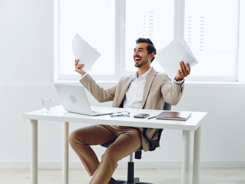 Lachende mannelijke ondernemer achter laptop met documenten, zichtbaar ontspannen en tevreden over zijn administratie.